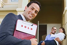 A realtor with a sold sign and a happy couple in the background
